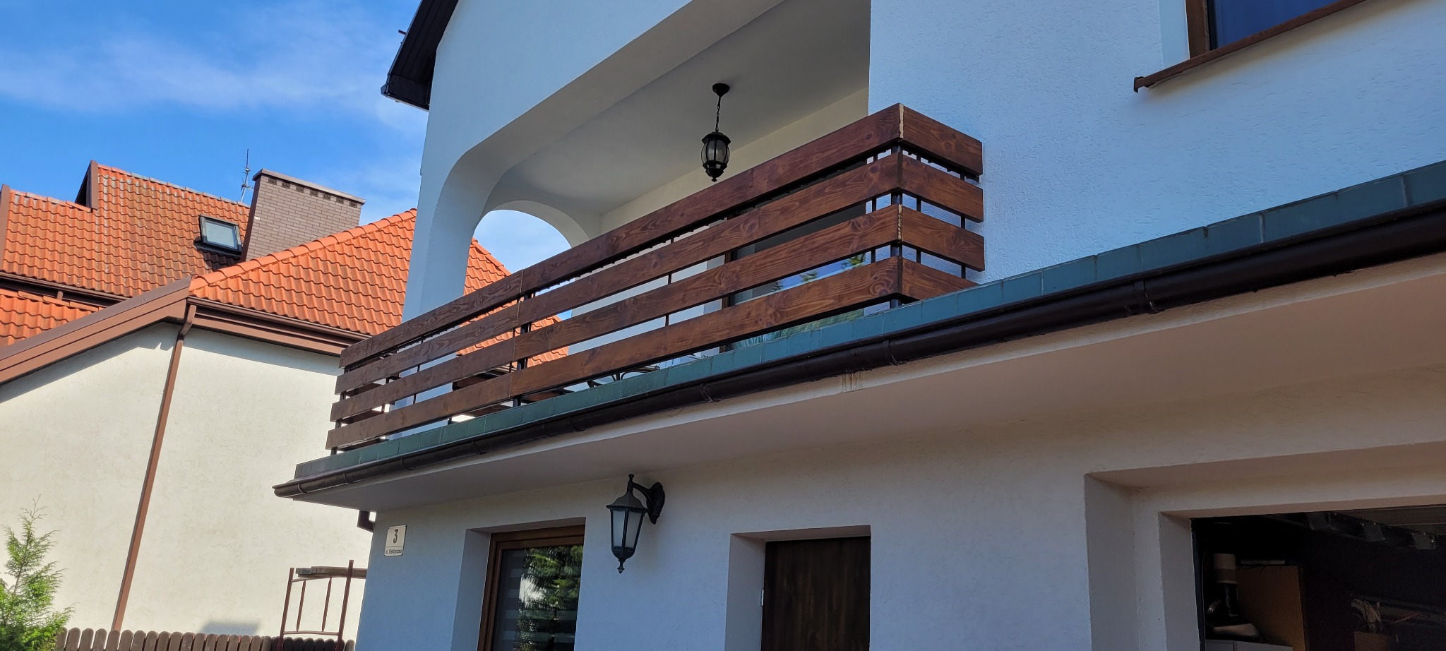 Balkon z drewnianą balustradą z poziomych desek, oświetlony wiszącą lampą w stylu retro, widok z dołu na biały budynek z elementami z czerwonej cegły w tle.