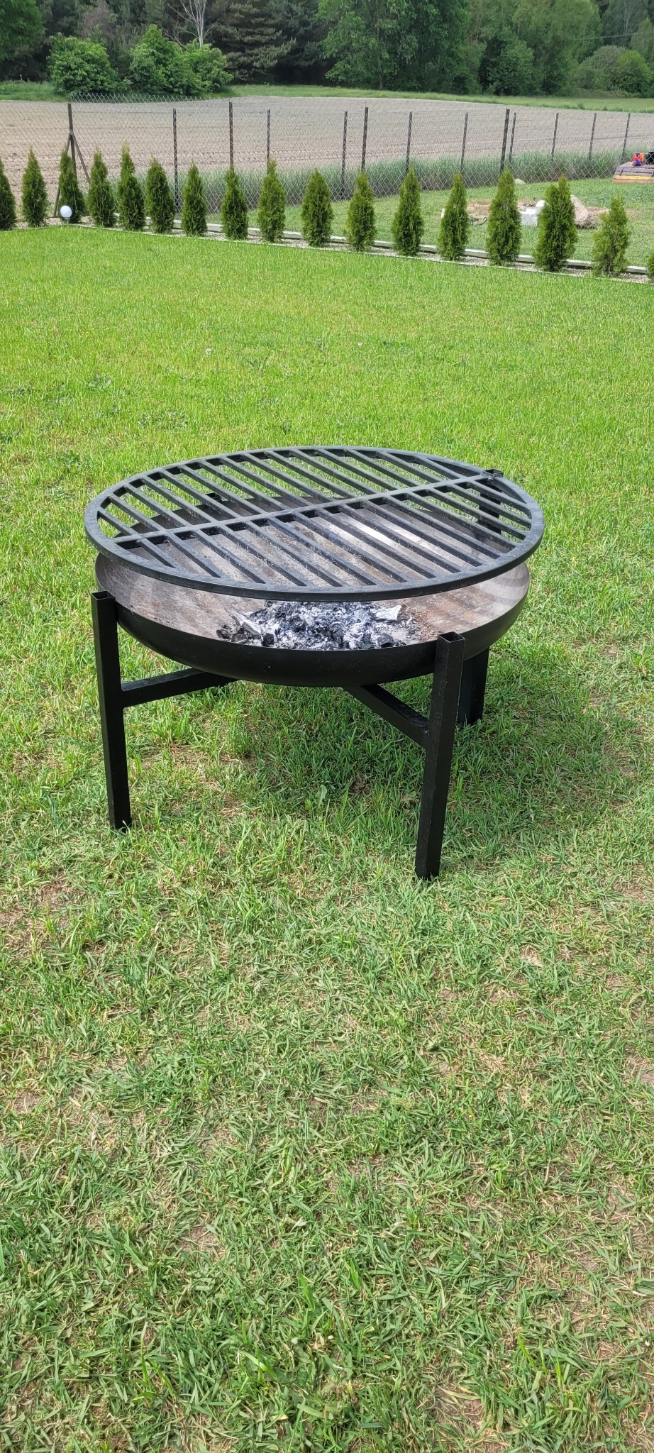 Metalowy grill ogrodowy z okrągłym rusztem i resztkami popiołu w palenisku, stojący na trawniku, w tle rząd tuj.