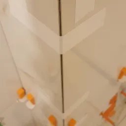 Narożnik ściany wyłożony białymi płytkami, zabezpieczony taśmą malarską i żółtą ścierką, z widocznymi pomarańczowymi krzyżykami dystansowymi i niebieską poziomicą w dolnej części kadru.