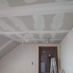 Poddasze z zamontowaną płytą kartonowo-gipsową, widoczne łączenia przed szpachlowaniem, przewody elektryczne zwisające z sufitu, drabina oparta o ścianę, widoczne profile mocujące.