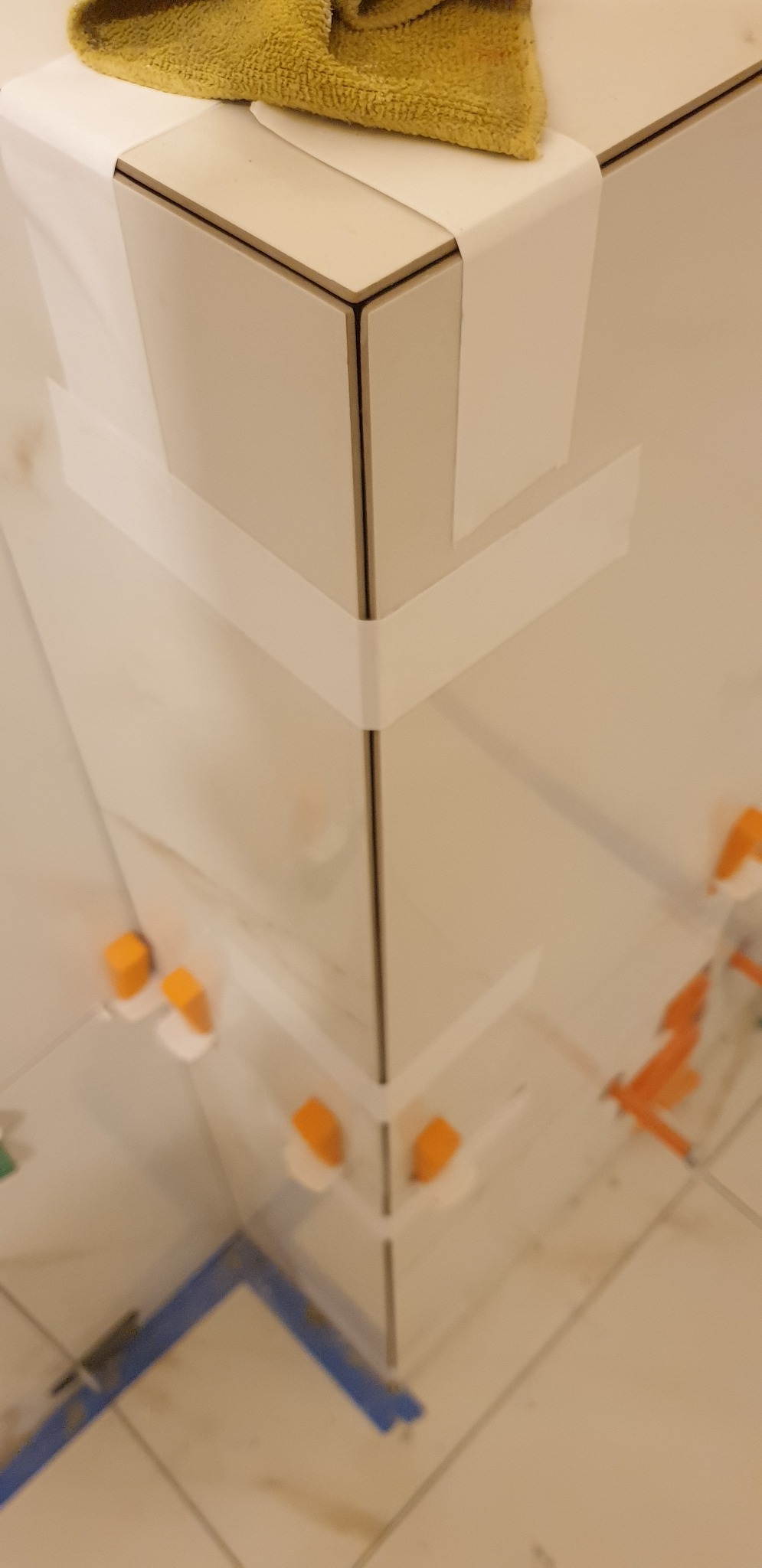 Narożnik ściany wyłożony białymi płytkami, zabezpieczony taśmą malarską i żółtą ścierką, z widocznymi pomarańczowymi krzyżykami dystansowymi i niebieską poziomicą w dolnej części kadru.