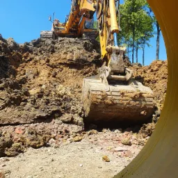 Żółta koparka na szczycie wykopu z widocznym operatorem, z boku fragment rury, w tle zieleń drzew i błękitne niebo.