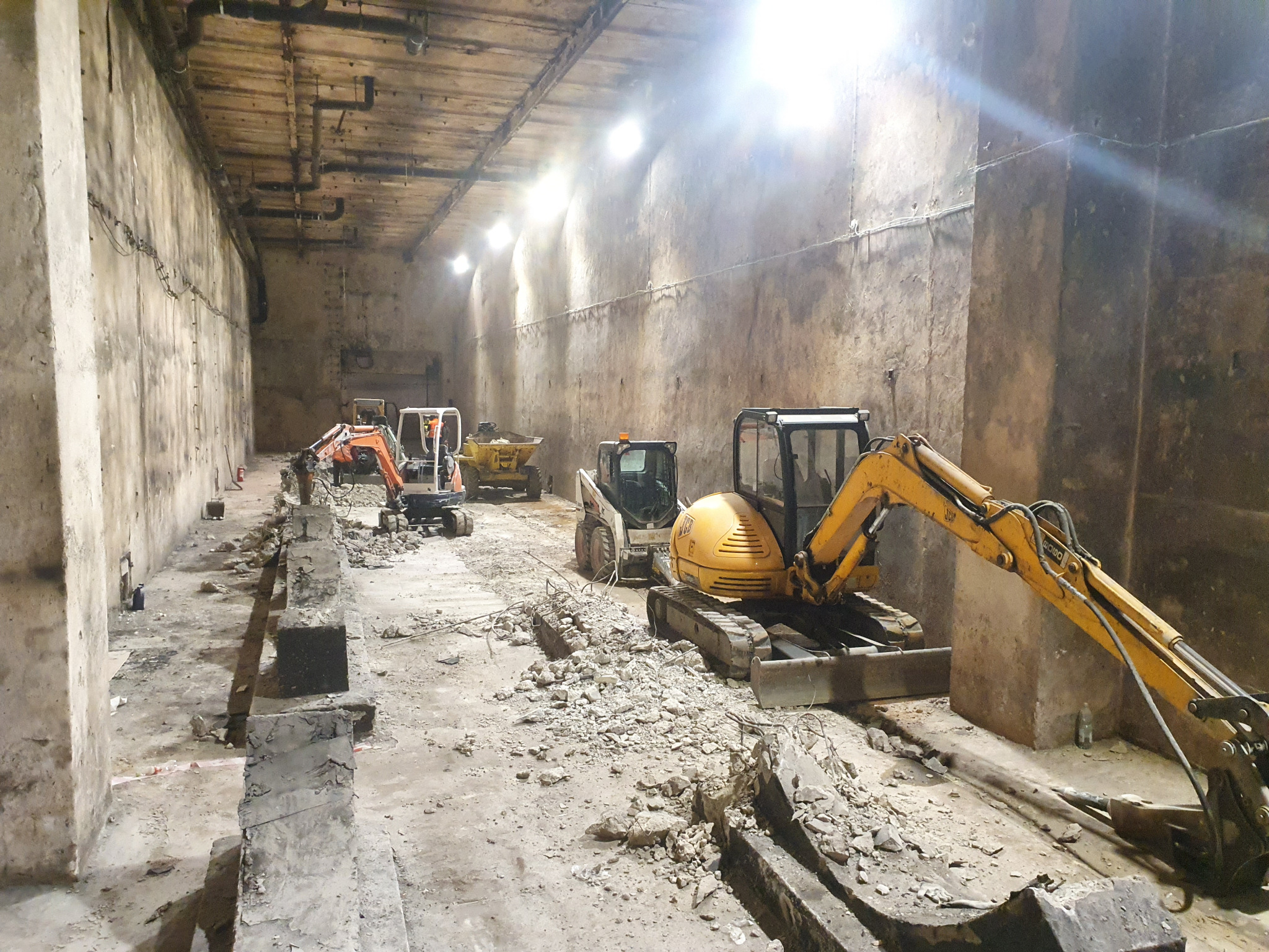Wnętrze hali z kilkoma maszynami budowlanymi, w tym żółtą koparką i białym bobcatem, pracującymi przy rozbiórce betonowej konstrukcji, widoczne gruzy i fragmenty zdemontowanych elementów.