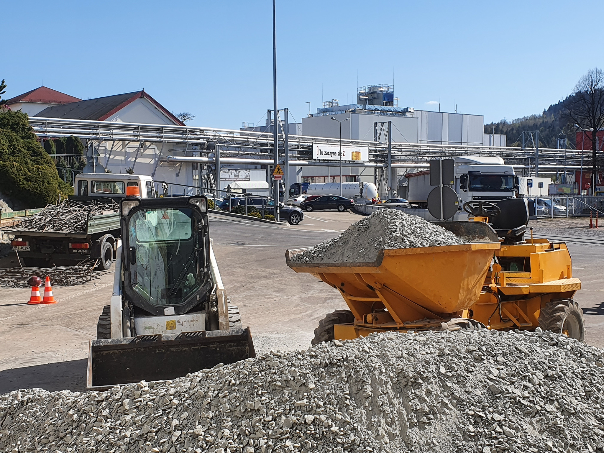 Żółta wywrotka i biała miniładowarka na placu budowy z hałdą gruzu, w tle budynek przemysłowy i ciężarówka MAN z ładunkiem metalowych prętów.