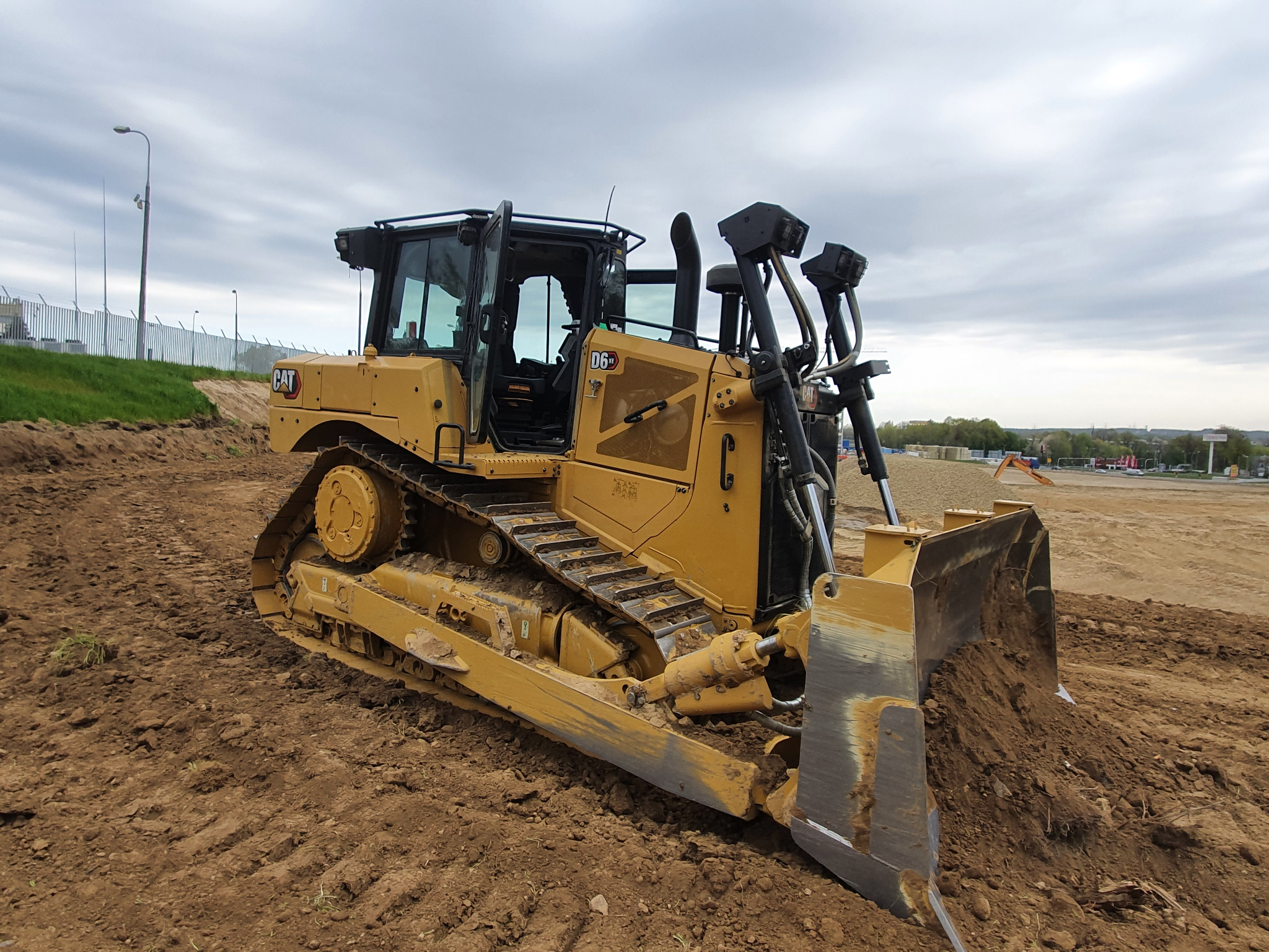Żółty spychacz marki CAT D6XE na placu budowy z otwartymi drzwiami kabiny, widoczne ślady gąsienic na ziemi, w tle ogrodzenie i pochmurne niebo.