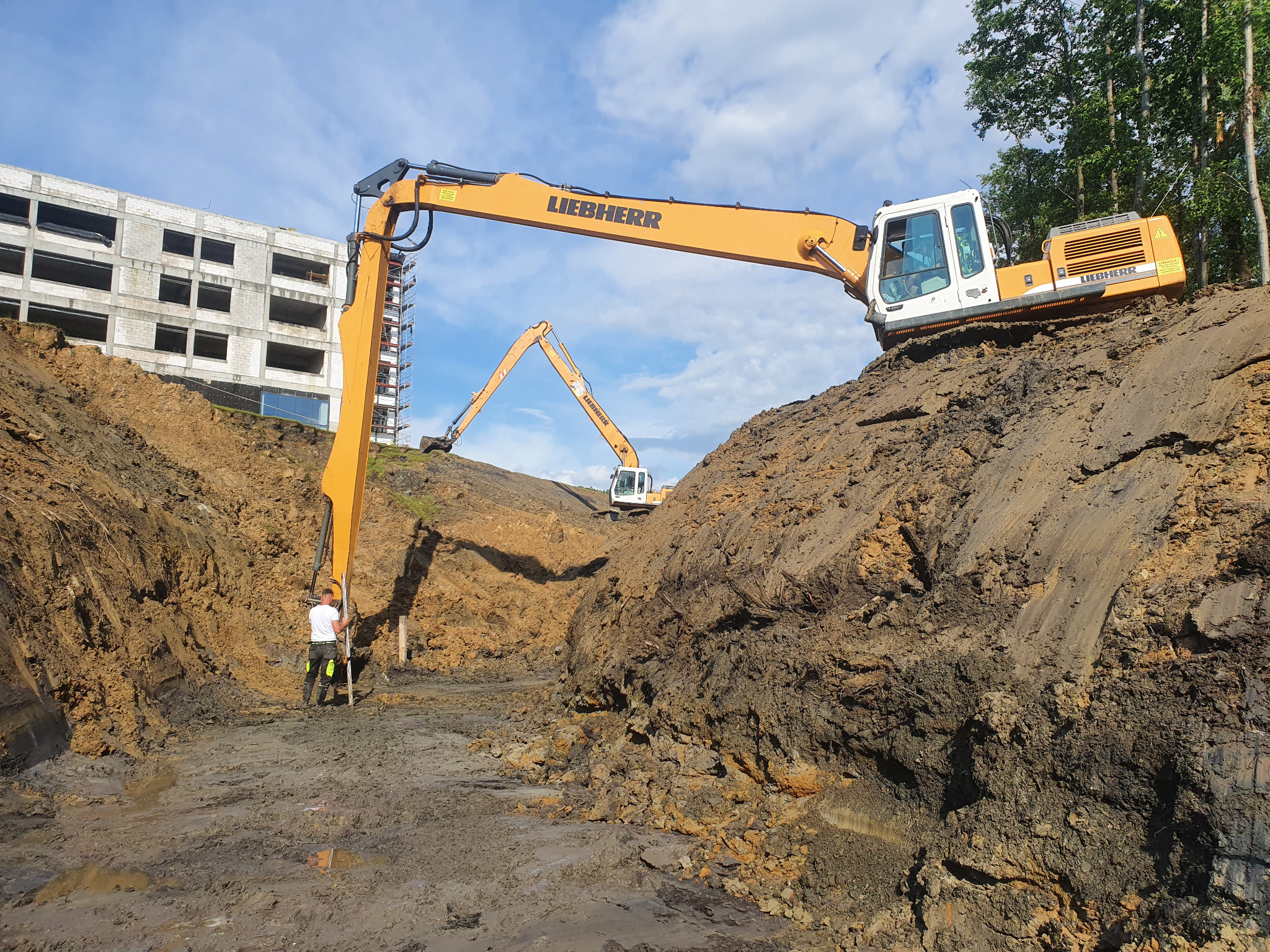 Żółty koparka Liebherr pracuje przy wykopie, w tle widoczny drugi pojazd i budynek w stanie surowym, pracownik z łatą mierniczą.