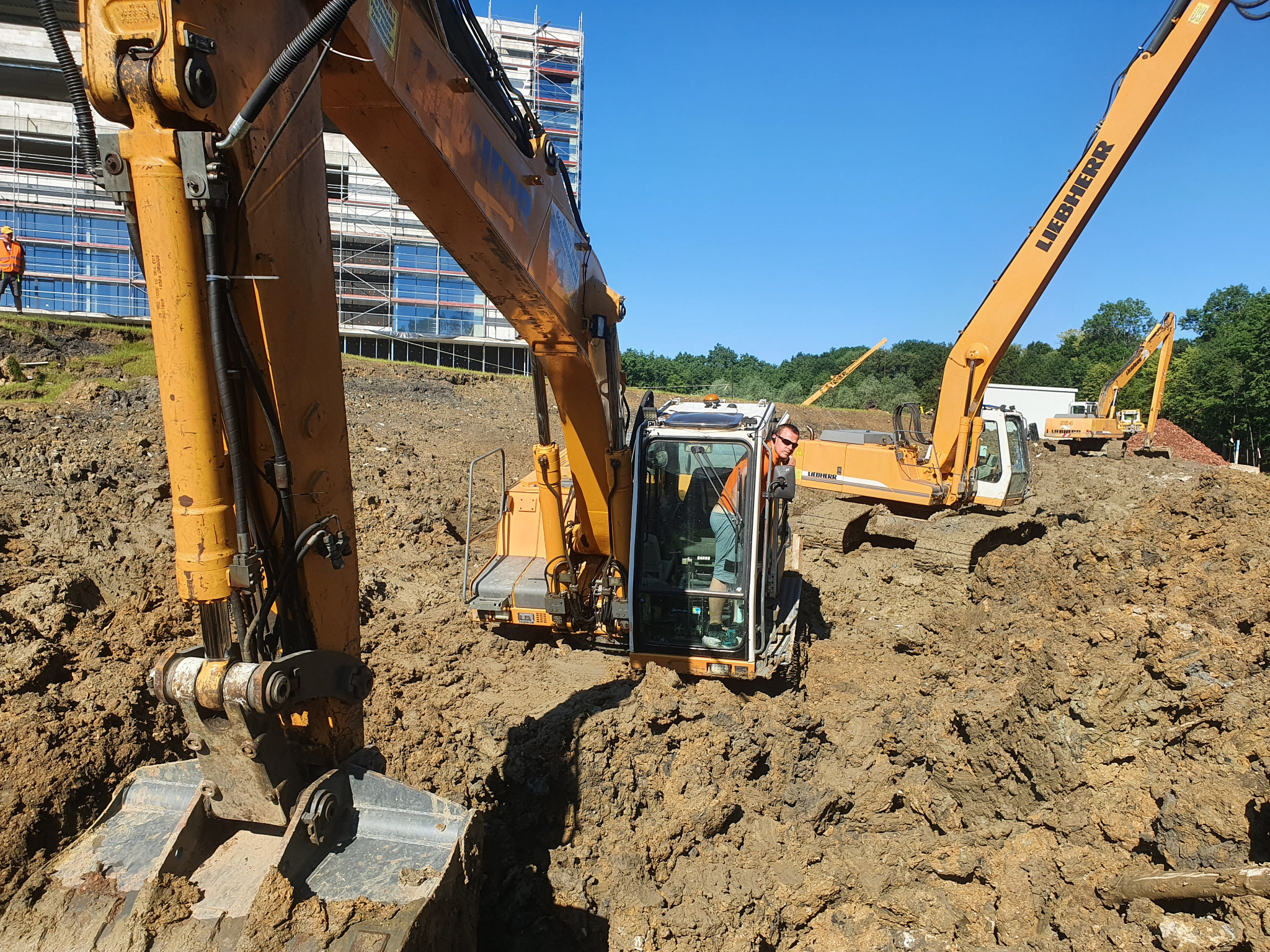 Żółta koparka Liebherr w trakcie prac ziemnych na budowie, operator w kabinie, w tle budynek w rusztowaniach i inne maszyny.