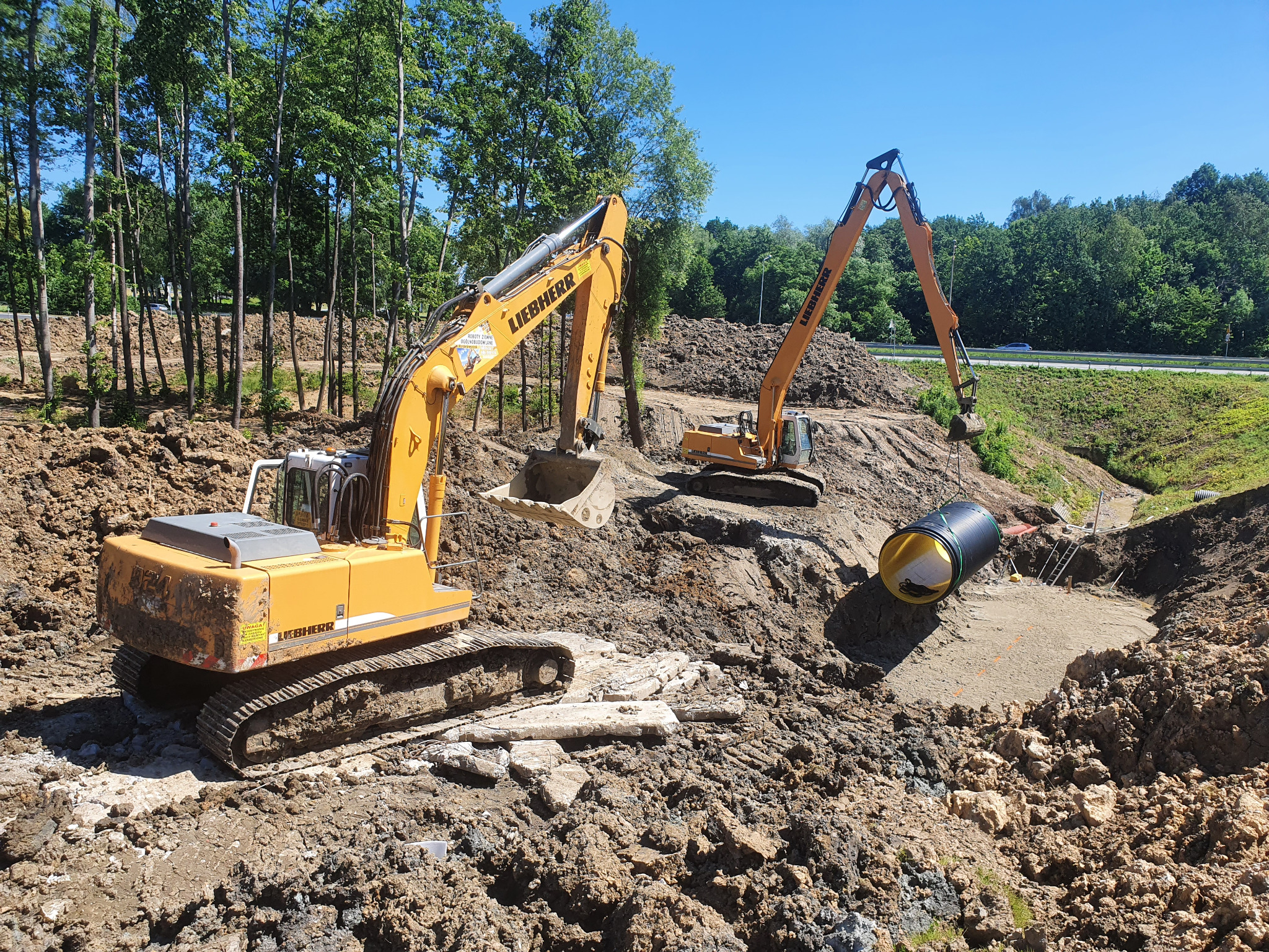 Dwa żółte koparki Liebherr pracują na placu budowy w słoneczny dzień, przygotowując wykop pod dużą, czarną rurę kanalizacyjną; w tle widoczny pas zieleni i droga.