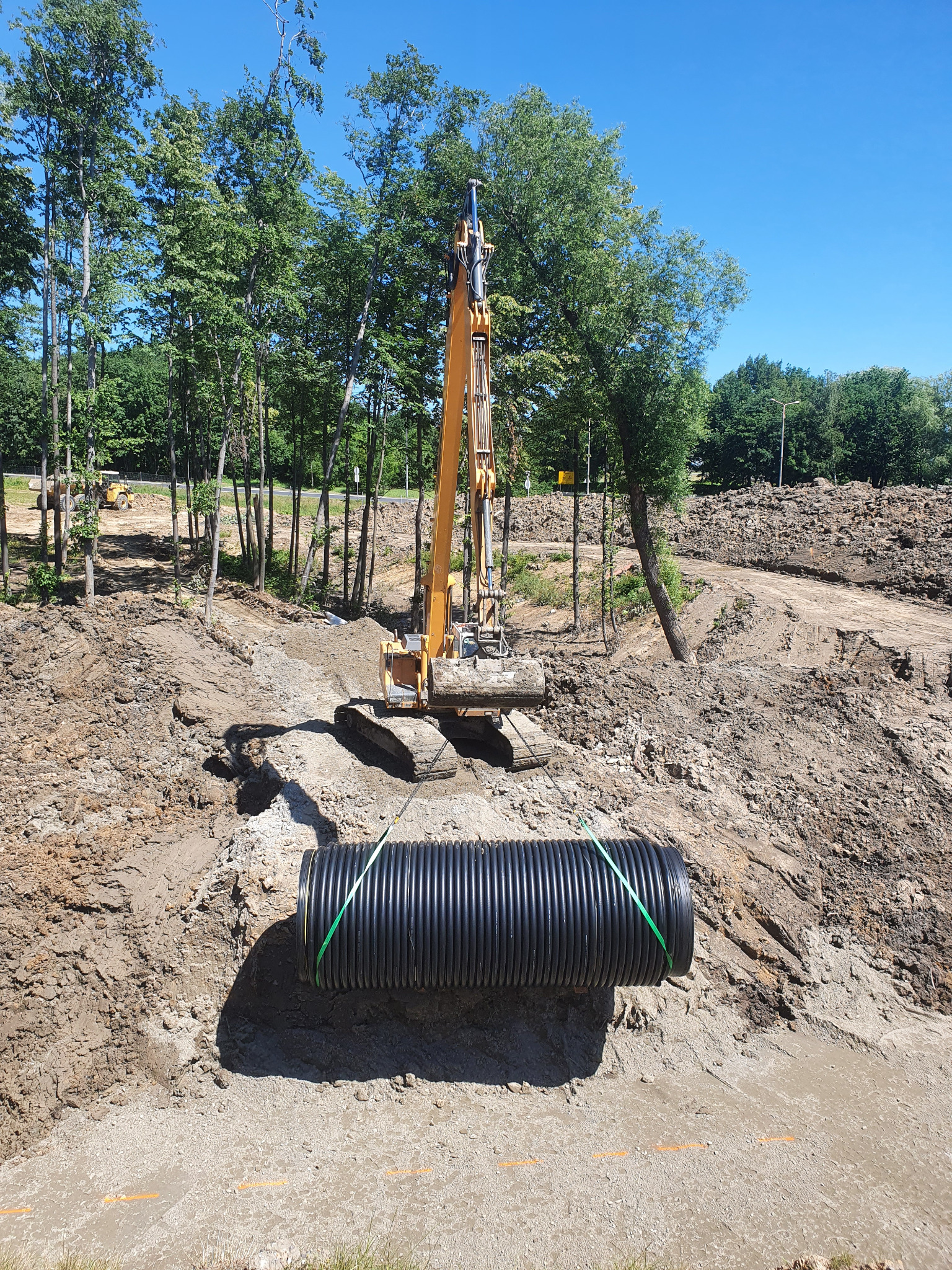 Żółty dźwig gąsienicowy umieszcza czarną rurę drenażową w wykopie na tle drzew i nieba.