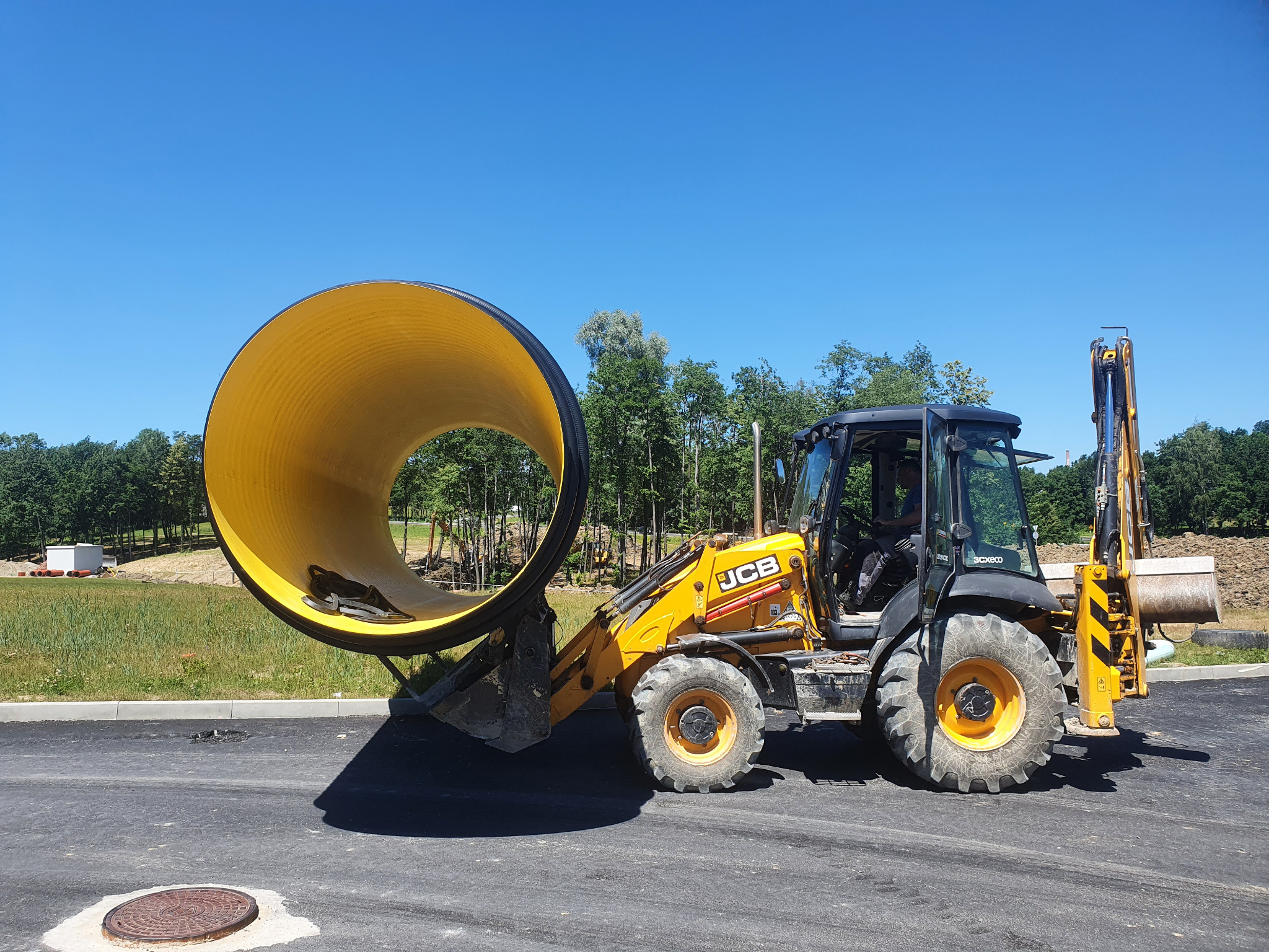 Żółta rura kanalizacyjna transportowana przez żółto-czarną koparko-ładowarkę JCB na tle zieleni i błękitnego nieba.