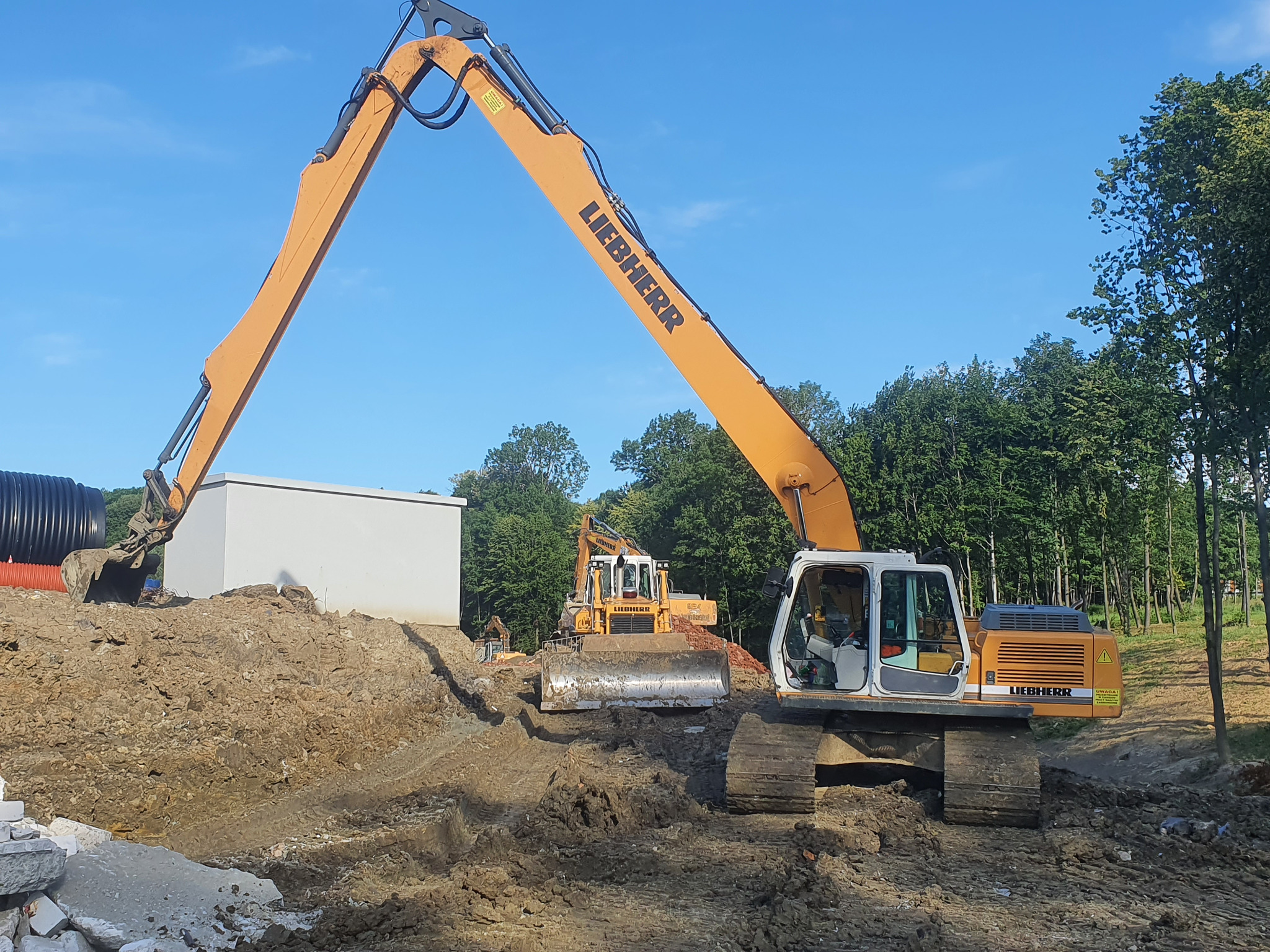 Żółta koparka Liebherr z uniesionym ramieniem i łyżką nad ziemią obok betonowego zbiornika, w tle widoczna druga koparka i rury kanalizacyjne.