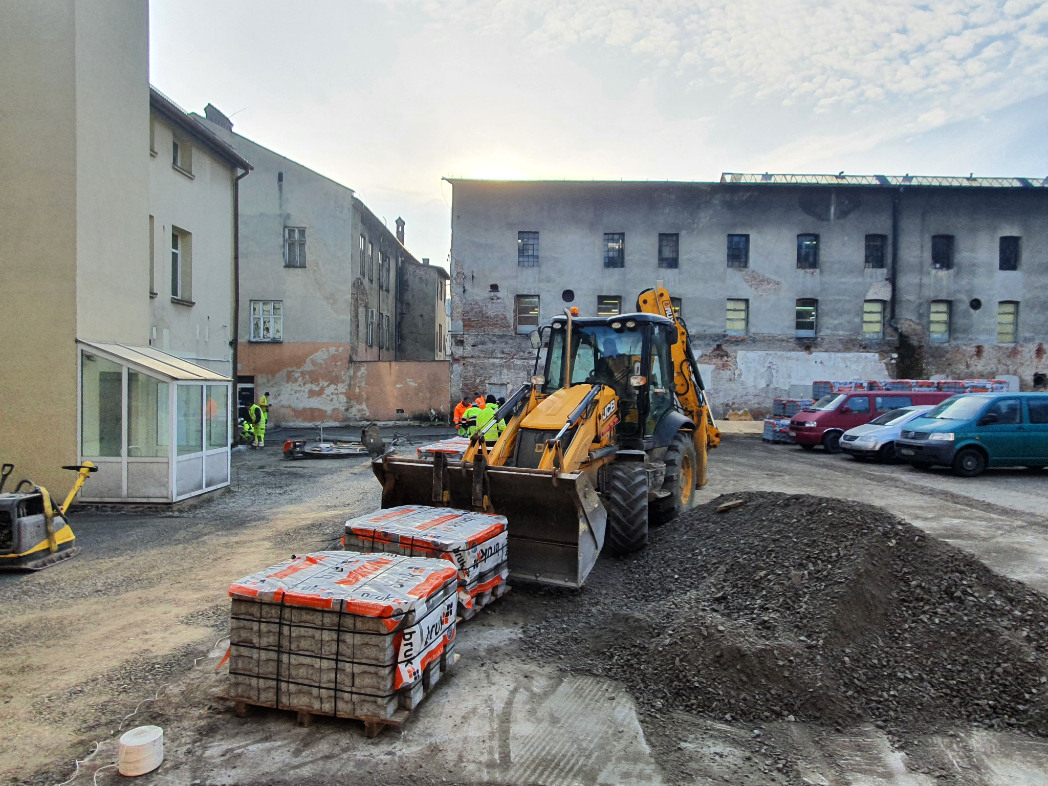 Żółta koparko-ładowarka JCB z dwoma paletami kostki brukowej na placu budowy, w tle budynki i pracownicy w odblaskowych kamizelkach.