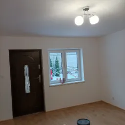 Świeżo pomalowane na biało wnętrze pokoju z brązowymi drzwiami wejściowymi i oknem z widokiem na zimowy krajobraz, widoczne akcesoria malarskie na podłodze.