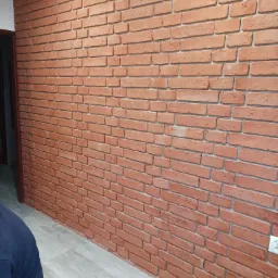 Ściana wewnętrzna wykończona dekoracyjną czerwoną cegłą, widoczny fragment podłogi z paneli imitujących drewno oraz fragment osoby w niebieskiej bluzie w lewym dolnym rogu.