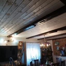 Drewniany sufit z widocznymi belkami w rustykalnym wnętrzu, widok z dołu na wykończony strop z boazerią, w tle meble i dekoracje.