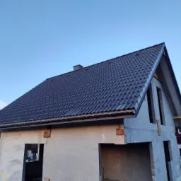 Niedokończony dom z ciemną dachówką ceramiczną, widoczne rynny i fragmenty niezabezpieczonych otworów okiennych, na tle błękitnego nieba.