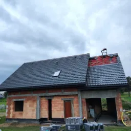 Dom w trakcie budowy, z częściowo położonym dachem z ciemnej dachówki, widoczne materiały budowlane marki Braas, fragment dachu w trakcie krycia z widoczną czerwoną membraną i rusztowaniem.