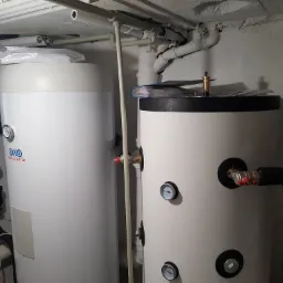 Dwa białe zbiorniki na wodę w piwnicy z widocznymi rurami i manometrami, jeden z logo producenta, a na drugim leżą dokumenty w niebieskiej teczce.