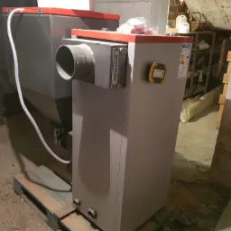 Dwa piece na paliwo stałe w szarej obudowie z czerwonymi akcentami, ustawione na palecie w piwnicy. Jeden z pieców posiada przyłącze do czyszczenia (WYCZYSTKA) oraz lampę roboczą. Widoczne...