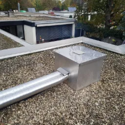 Instalacja wentylacyjna z dwoma elastycznymi przewodami aluminiowymi, wychodząca z metalowej skrzynki na płaskim dachu pokrytym drobnymi kamieniami, w tle budynki z zielonymi dachami i drzewa.