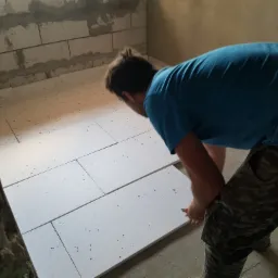 Instalacja białych płyt izolacyjnych na podłodze w surowym wnętrzu, pracownik w niebieskiej koszulce i spodniach moro przycina płytę nożykiem.