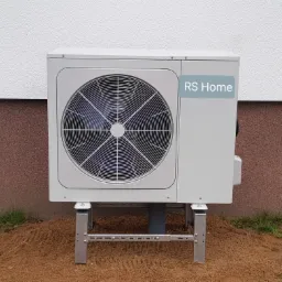 Zewnętrzna jednostka klimatyzacji z naklejką RS Home, zamontowana na stalowej konstrukcji przy ścianie budynku z widocznym fragmentem trawnika.