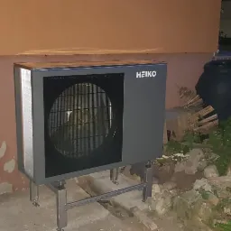 Szara pompa ciepła marki HEIKO zamontowana na metalowej konstrukcji, widoczna przy ścianie budynku z oknem i elementami dekoracyjnymi ogrodu.