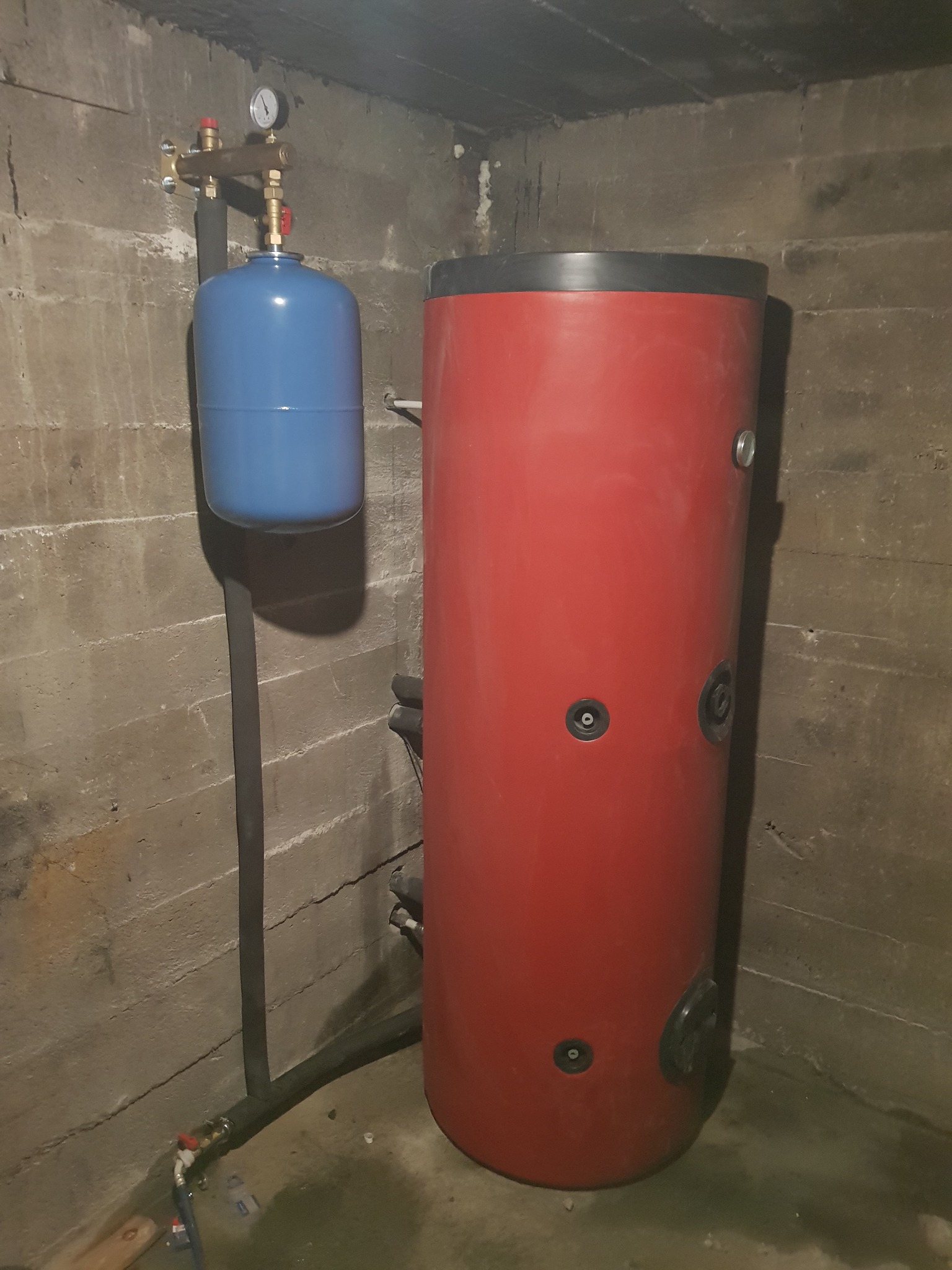 Czerwony zasobnik ciepłej wody użytkowej i niebieskie naczynie przeponowe z manometrem, zamontowane w surowym pomieszczeniu z betonowymi ścianami i podłogą.