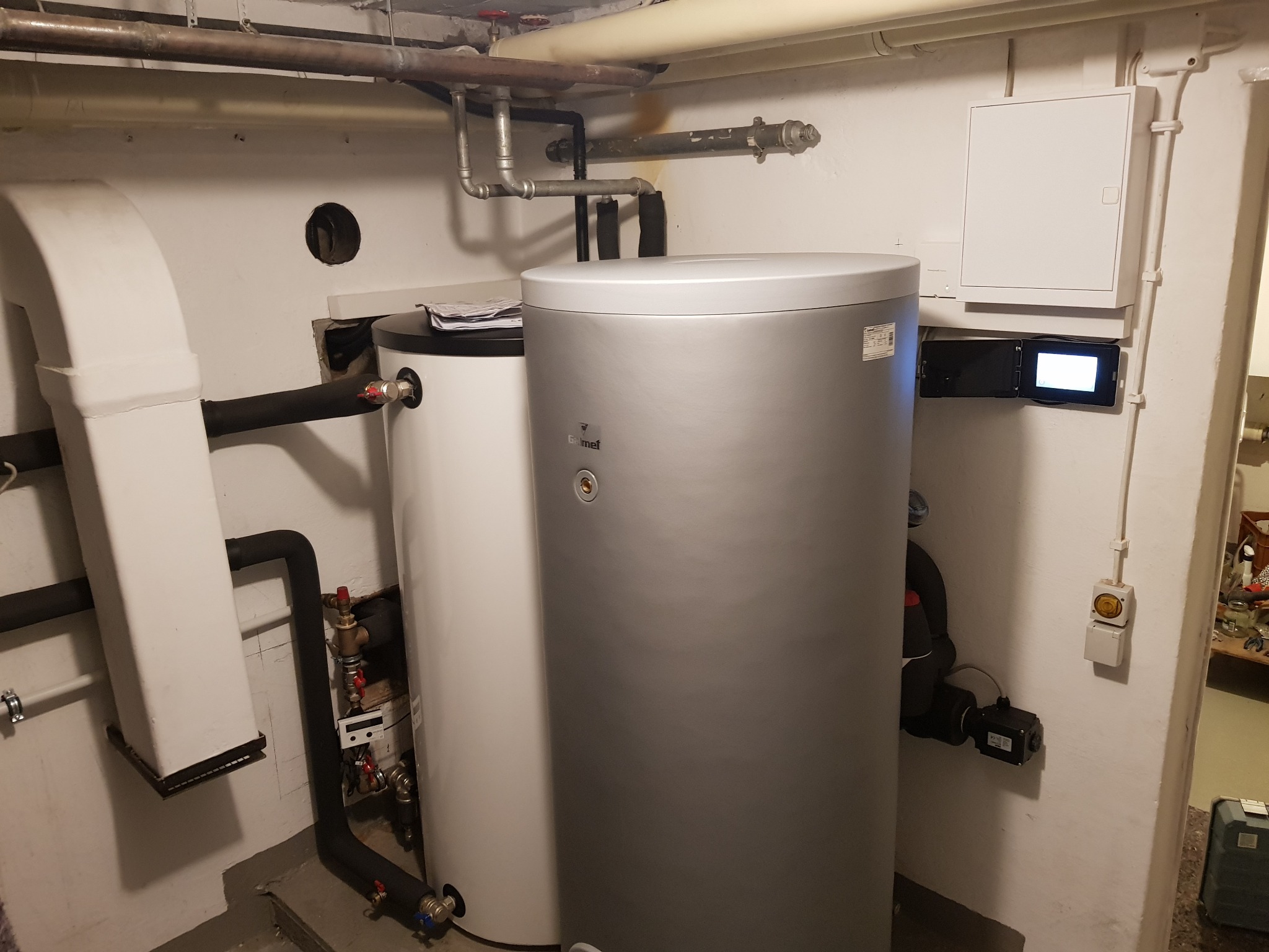 Dwa zbiorniki akumulacyjne ciepła w kotłowni, jeden biały, drugi srebrny, z widocznymi rurami i zaworami, na ścianie skrzynka elektryczna i panel sterowania z wyświetlaczem.