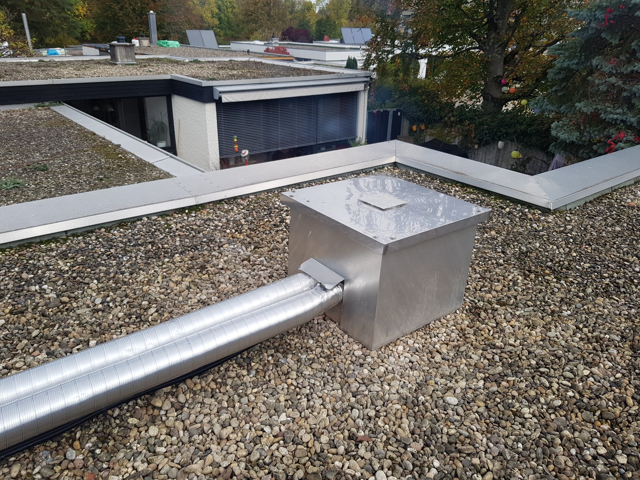 Instalacja wentylacyjna z dwoma elastycznymi przewodami aluminiowymi, wychodząca z metalowej skrzynki na płaskim dachu pokrytym drobnymi kamieniami, w tle budynki z zielonymi dachami i drzewa.