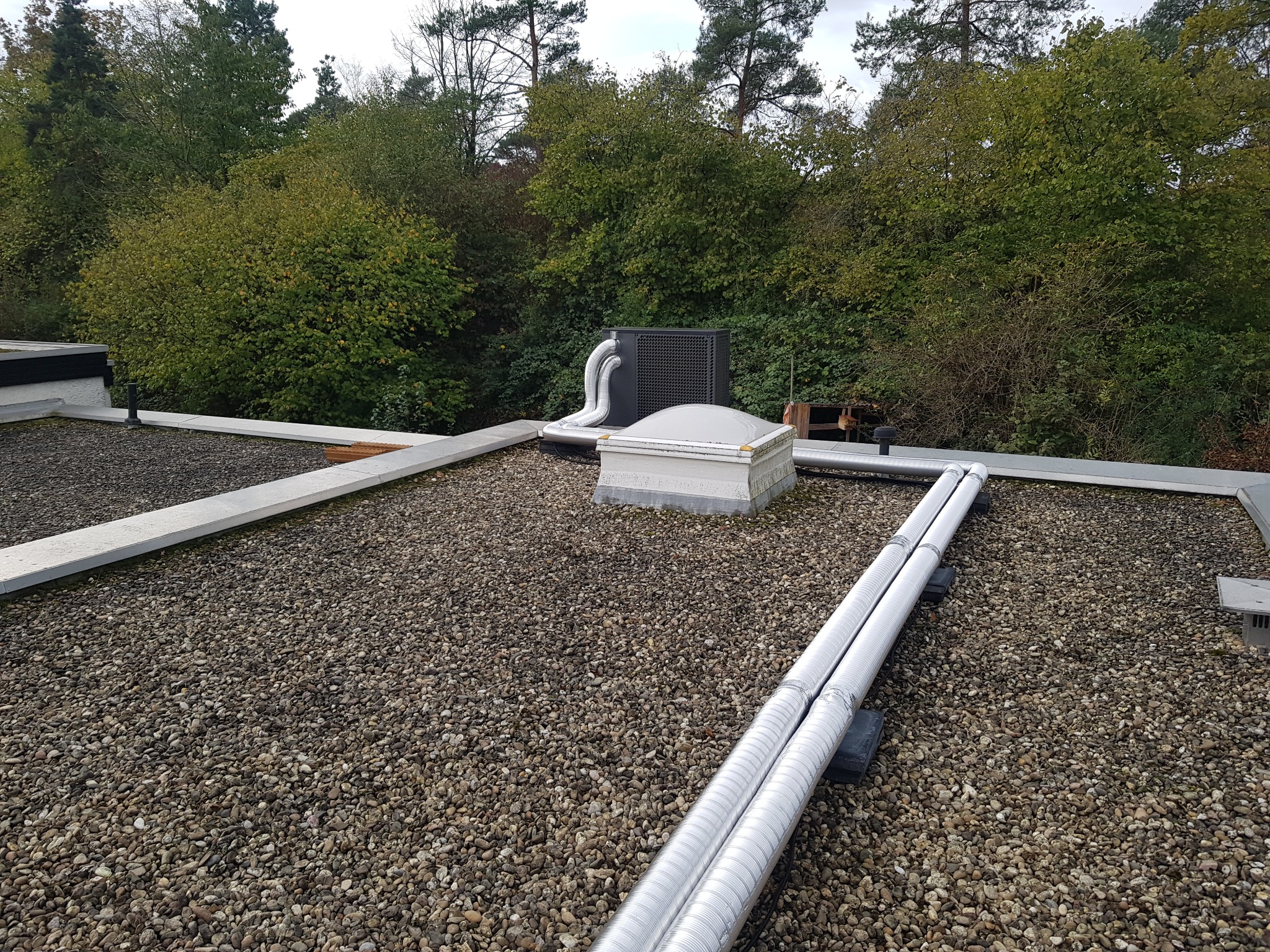 Czarna jednostka zewnętrzna z dwoma rurami na płaskim dachu pokrytym żwirem, widoczny świetlik dachowy i zieleń w tle.