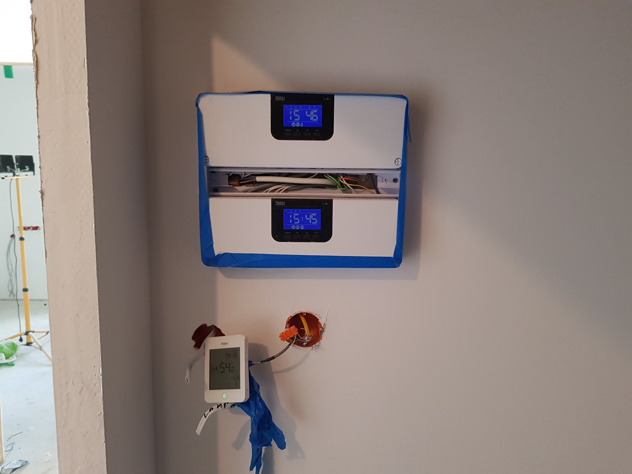 Dwa regulatory temperatury TECH L-9 zamontowane jeden nad drugim na ścianie, zabezpieczone niebieską taśmą malarską, z podłączonymi przewodami. Obok widoczny termostat pokojowy Haier z wyświetlaną...