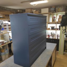 JKMAK Jarosław Mąkosa - Niebieska komoda z szufladami w warsztacie, widoczne narzędzia i pojemniki z farbą, surowe oświetlenie.