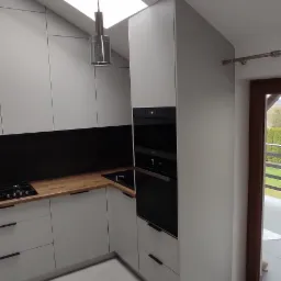 Jasnoszara zabudowa kuchenna z drewnianym blatem, czarnym panelem nad blatem, piekarnikiem w zabudowie, oświetlona światłem z okna dachowego i nowoczesną lampą wiszącą.