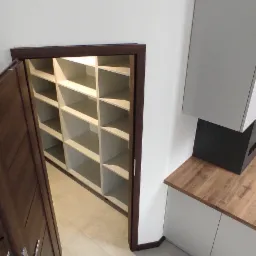 Wnętrze z otwartą, brązową drewnianą szafą wnękową z półkami, widok z góry na jasną podłogę i fragment zabudowy kuchennej z drewnianym blatem.