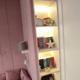 Biała, podświetlana p&oacute;łka wbudowana w ścianę obok ł&oacute;żka z r&oacute;żowym zagł&oacute;wkiem, na p&oacute;łkach książki, ozdoby i dekoracje.