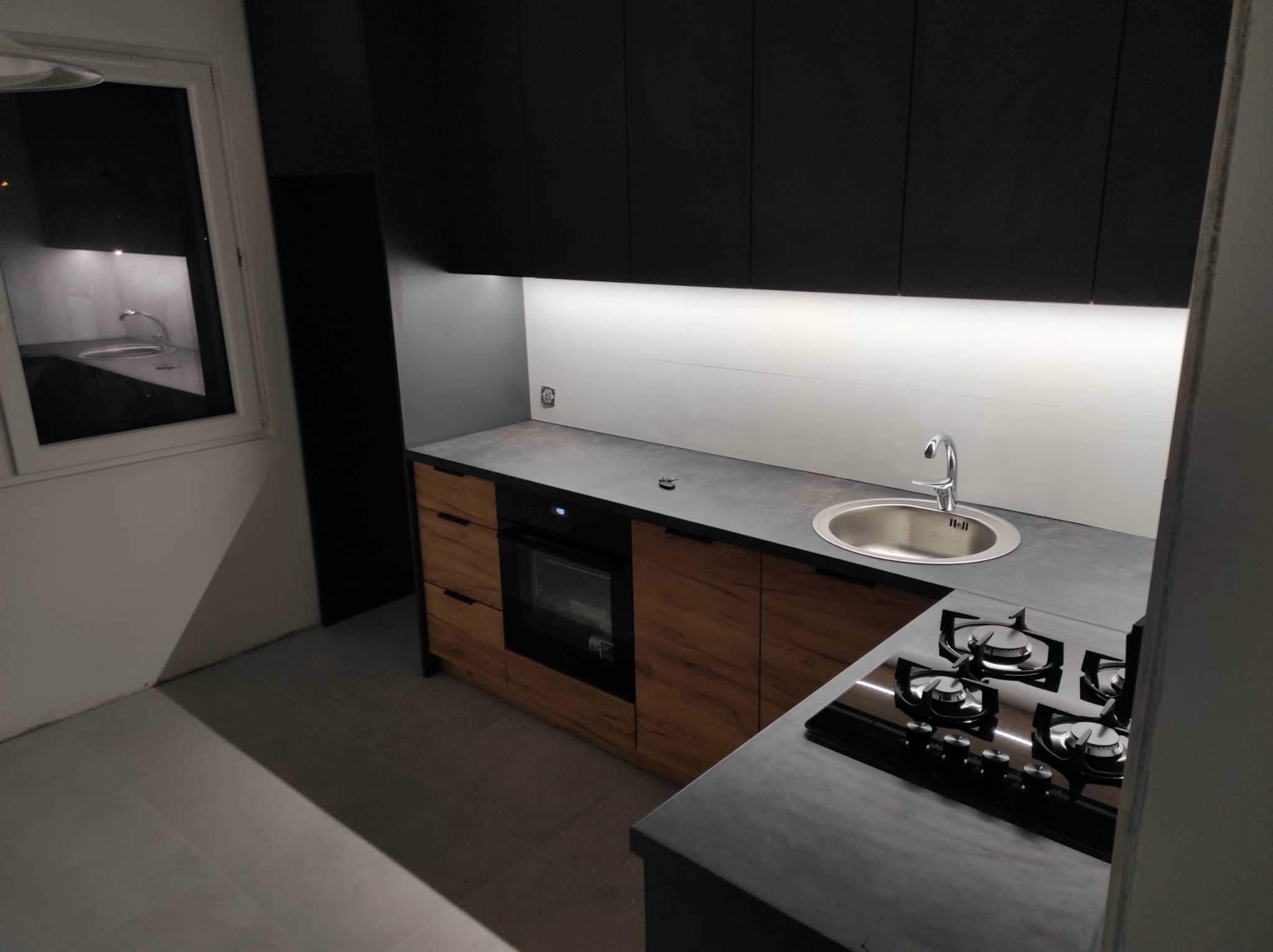 Nowoczesna kuchnia: czarne szafki górne, drewnopodobne szafki dolne, szary blat, płyta gazowa z czarnego szkła i oświetlenie LED pod szafkami.