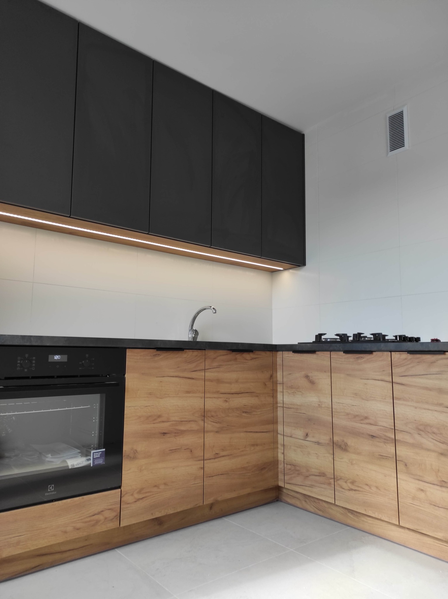 Nowoczesna kuchnia z drewnianymi szafkami dolnymi i czarnymi górnymi, podświetlona listwą LED, z czarnym blatem i sprzętem AGD Electrolux.