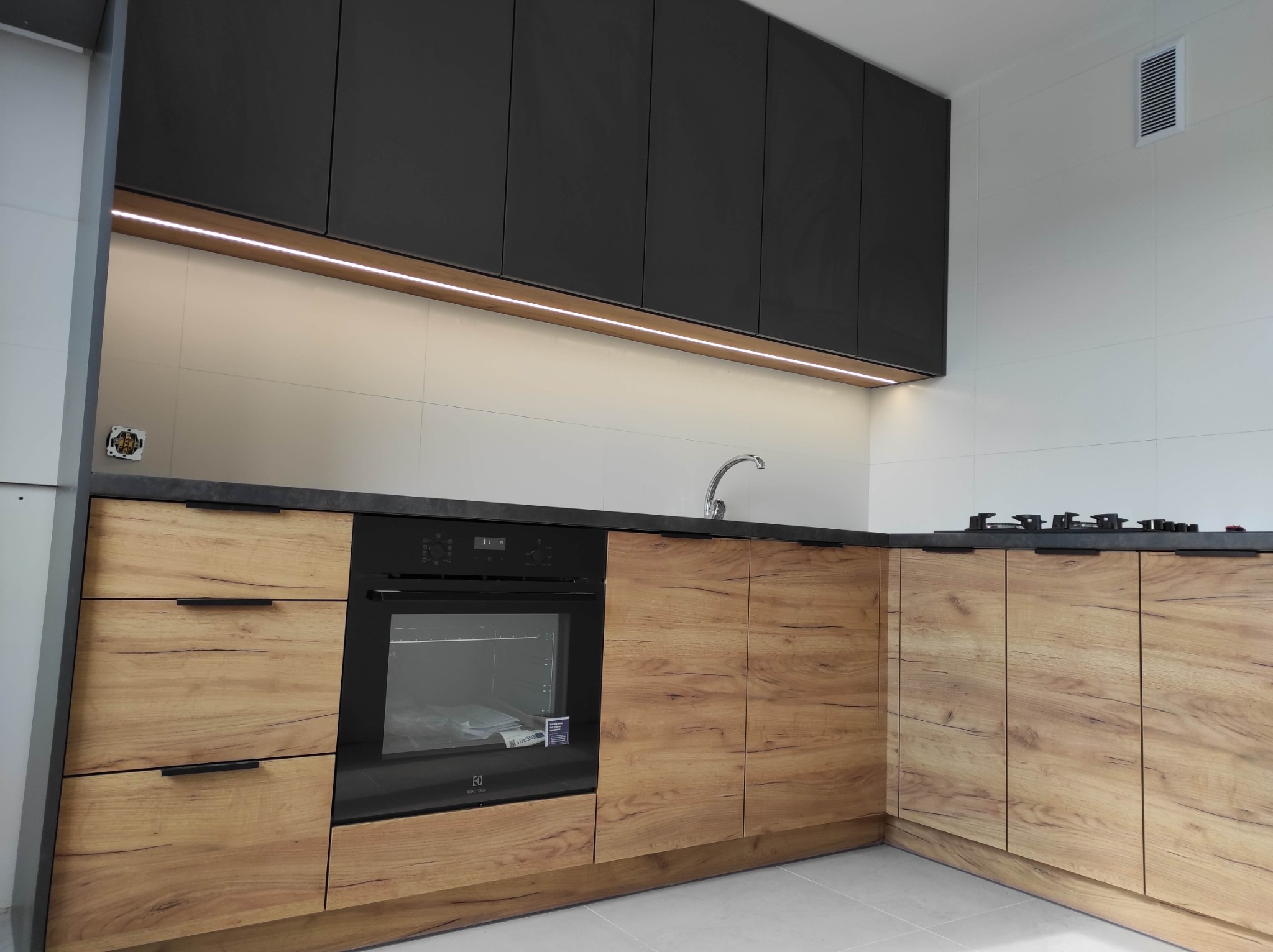 Nowoczesna kuchnia z drewnianymi frontami szafek dolnych i czarnymi szafkami górnymi, oświetlenie LED pod szafkami, płyta grzewcza i piekarnik w zabudowie, minimalistyczny zlew.