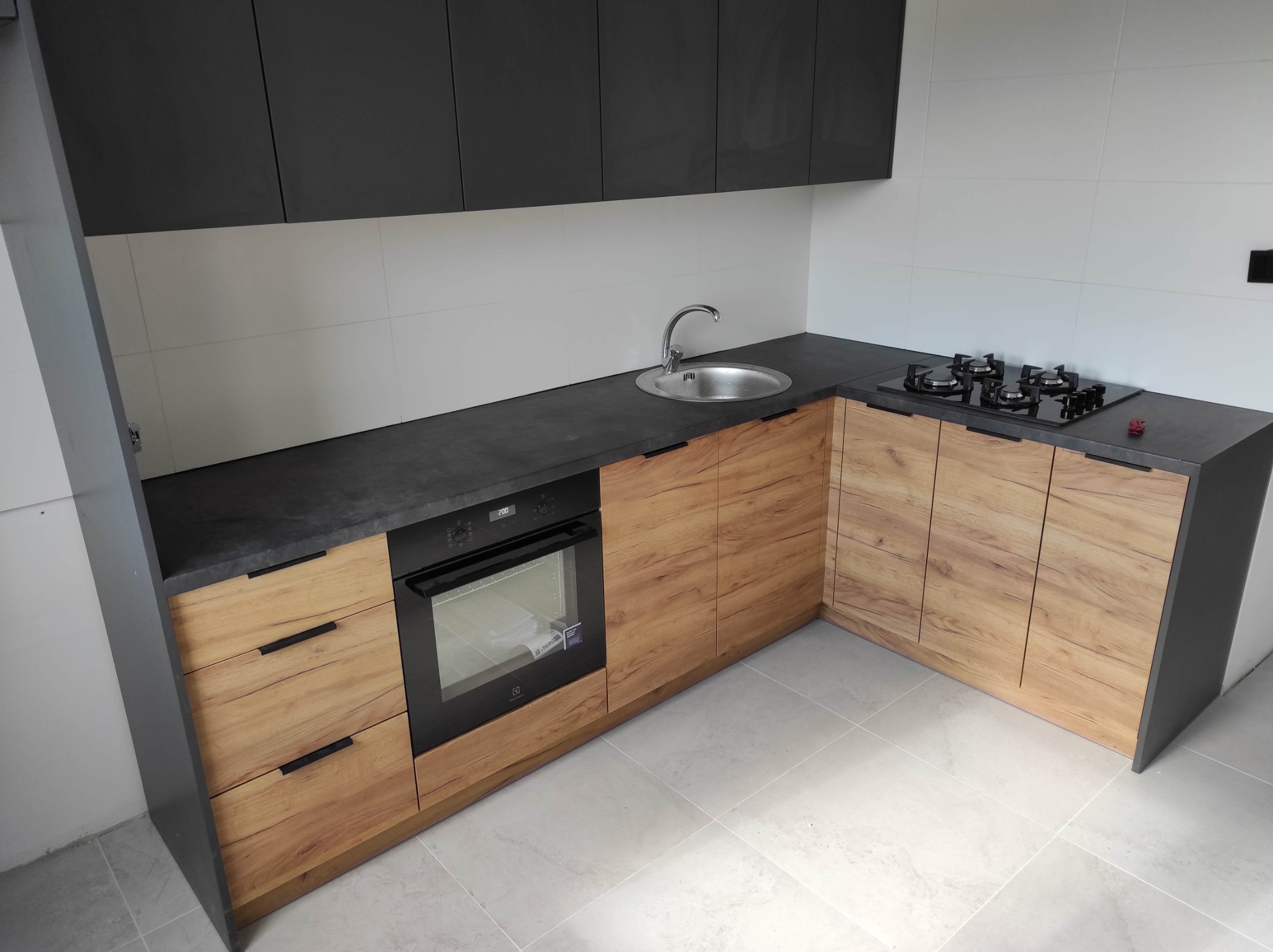 Nowoczesna kuchnia z drewnianymi frontami szafek dolnych i grafitowymi szafkami górnymi, czarny blat z wbudowanym zlewem i płytą gazową, piekarnik Electrolux w zabudowie.