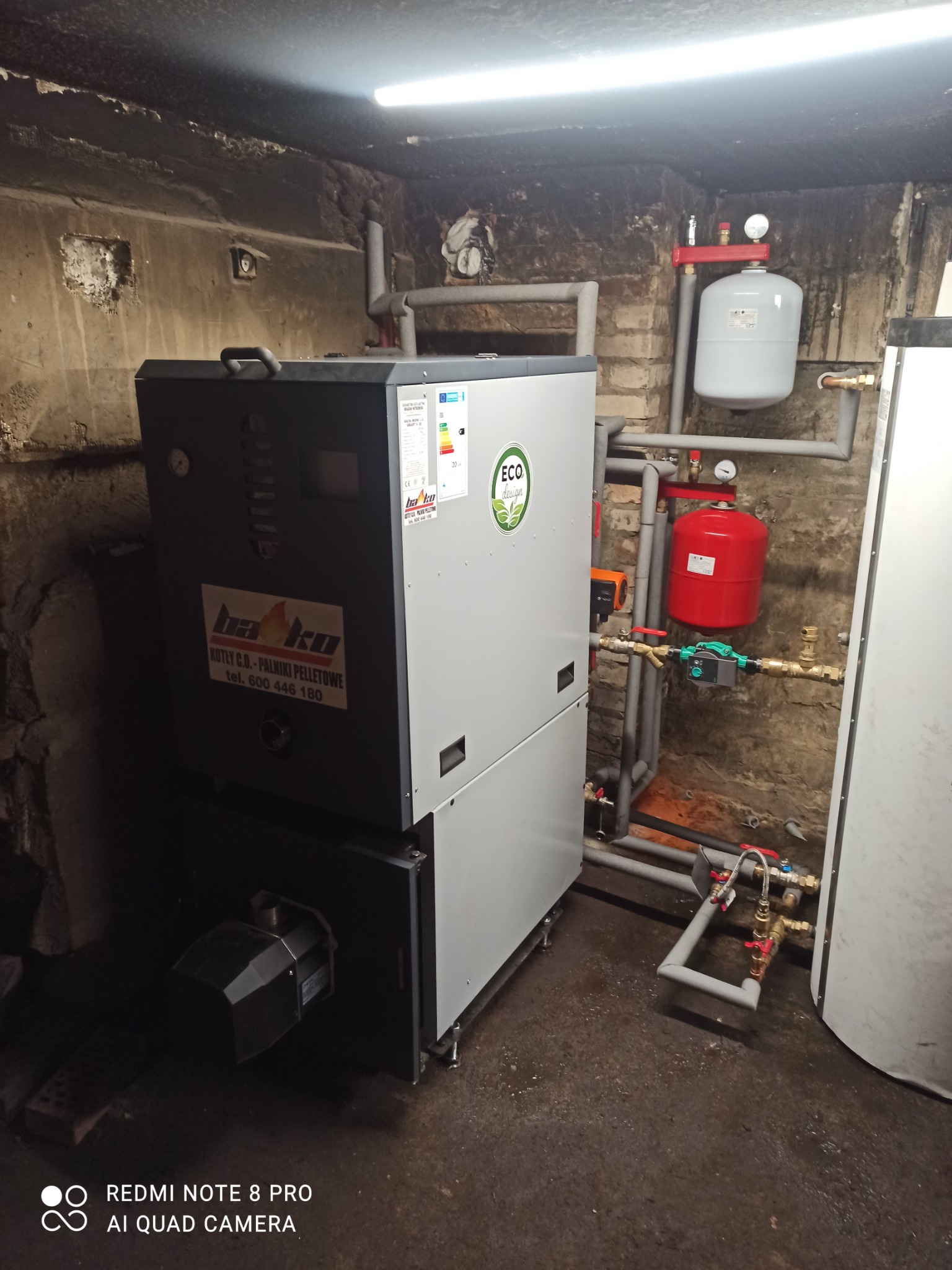 Kotłownia z zainstalowanym kotłem na pellet firmy BaKo, widoczne rury, zbiorniki wyrównawcze, ściany z surowego betonu i cegły.