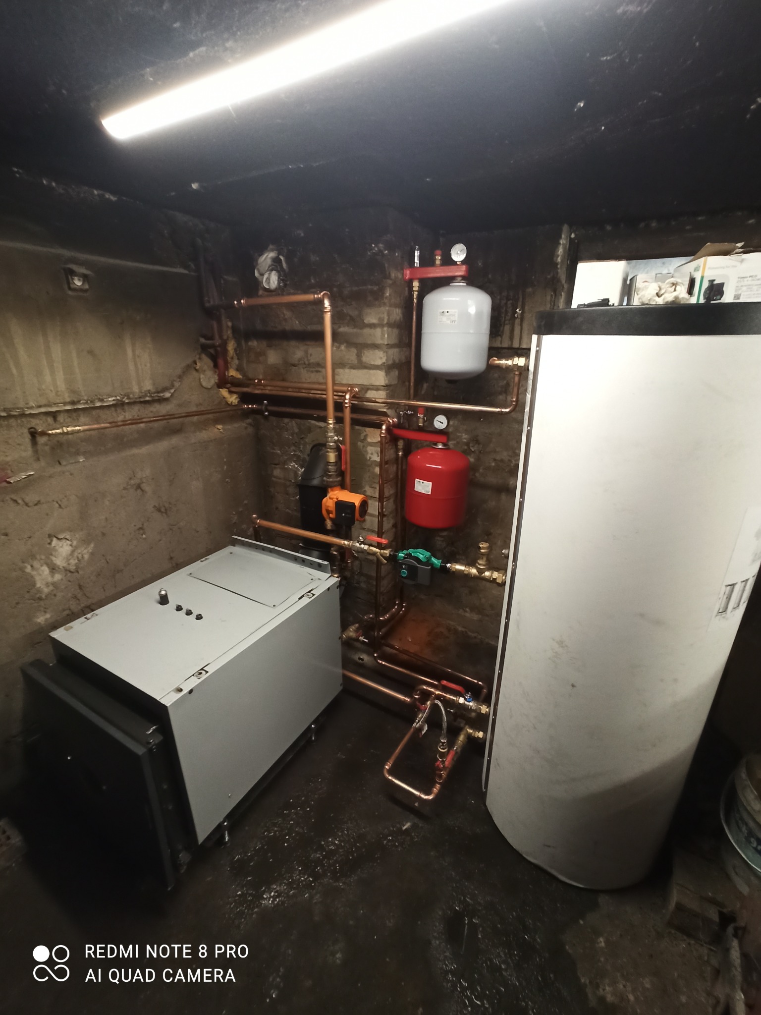 Zainstalowany system ogrzewania w piwnicy: szary piec obok białego zasobnika, czerwony zbiornik wyrównawczy, miedziane rury i osprzęt.