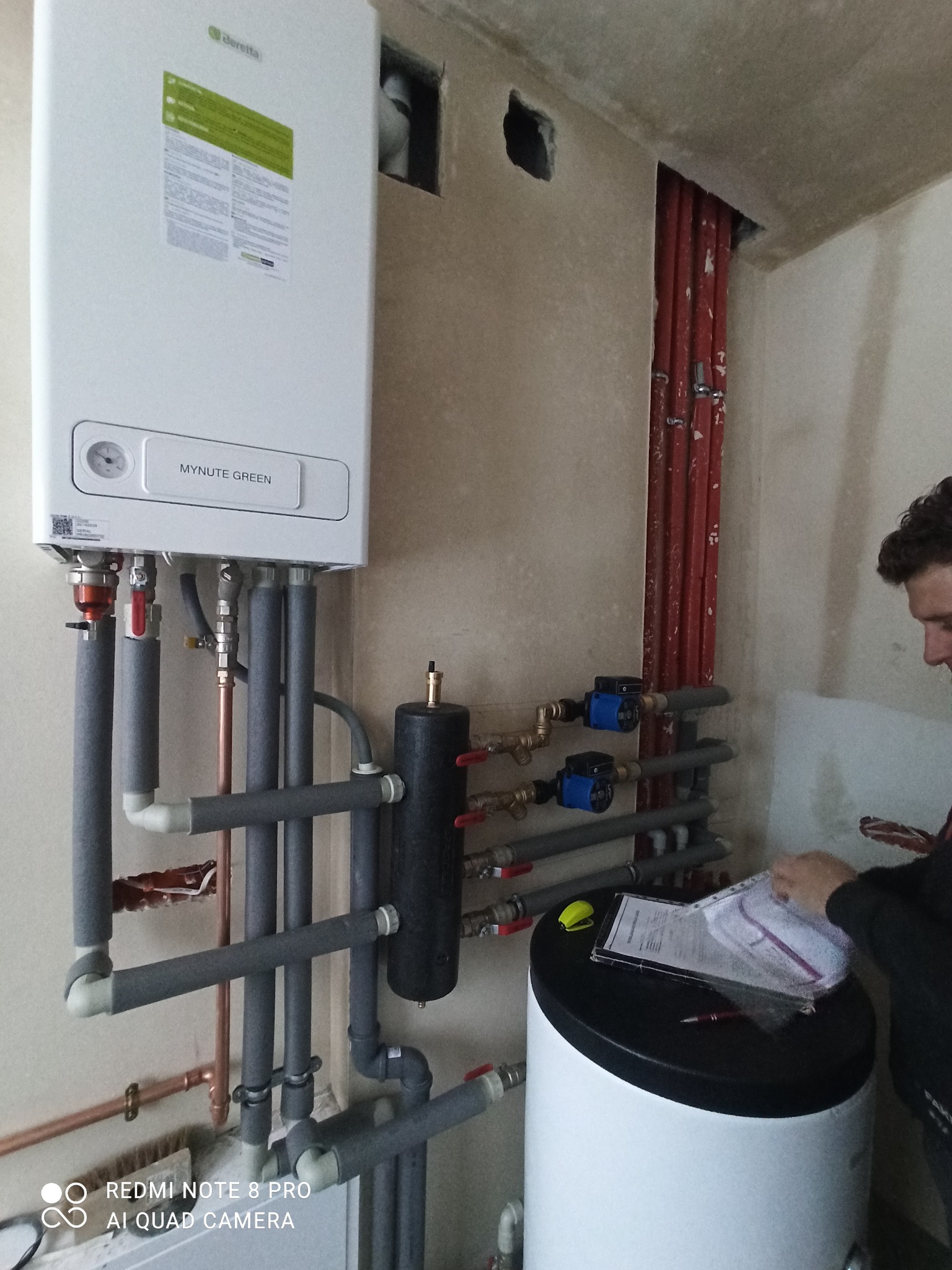 Biały wiszący piec gazowy Beretta Mynute Green z podłączonymi rurami, zbiornik na wodę i instalator przeglądający dokumenty.