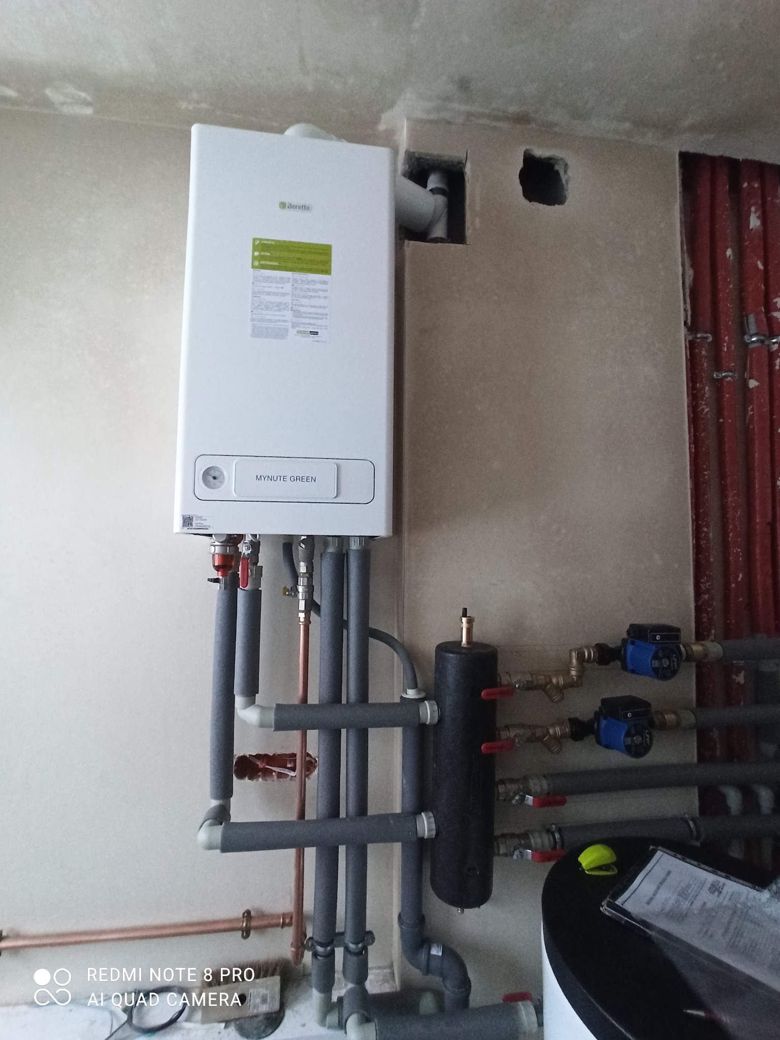 Zamontowany, biały piec gazowy Beretta Mynute Green z orurowaniem, widoczne pompy obiegowe, instalacja w surowym stanie, ściany w trakcie wykańczania.