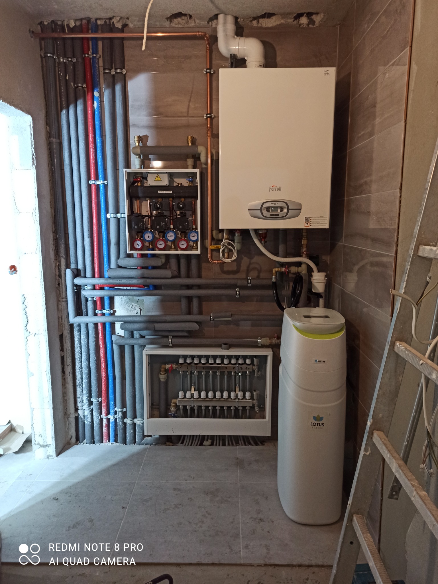 Kotłownia w trakcie montażu: biały piec gazowy Ferolli, rozdzielacz z manometrami, rury w izolacji, zmiękczacz wody Lotus System i drabina oparta o ścianę.