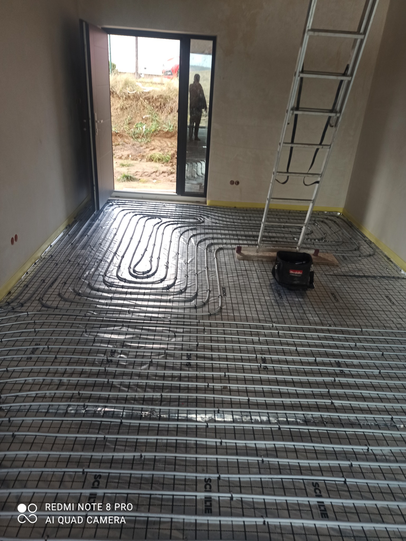 Rozłożona instalacja ogrzewania podłogowego na siatce w surowym wnętrzu, widoczna drabina i torba narzędziowa, zarys postaci w otwartych drzwiach.