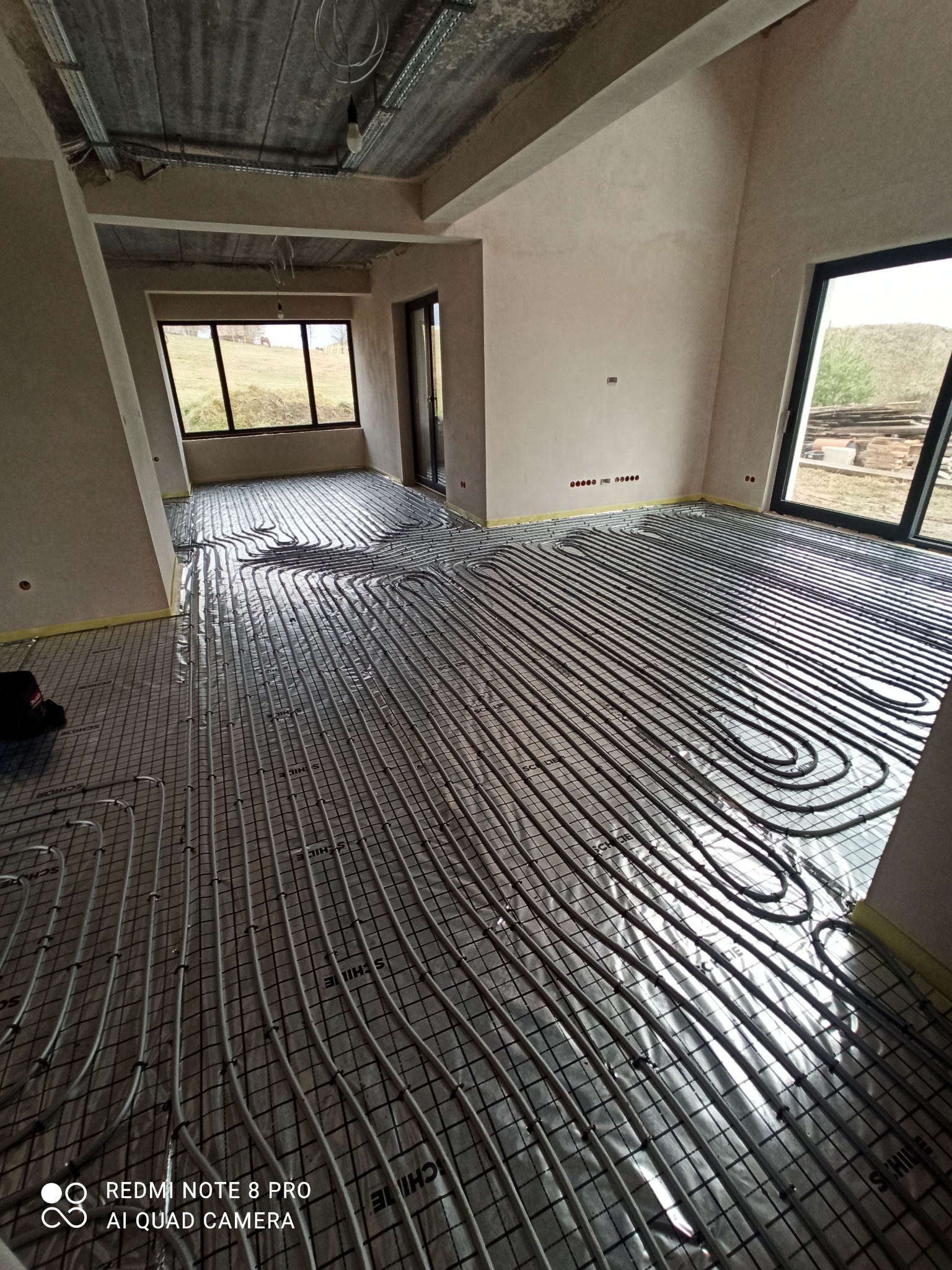 Szare rury ogrzewania podłogowego ułożone na srebrnej folii z nadrukiem w surowym wnętrzu z oknami wychodzącymi na krajobraz.