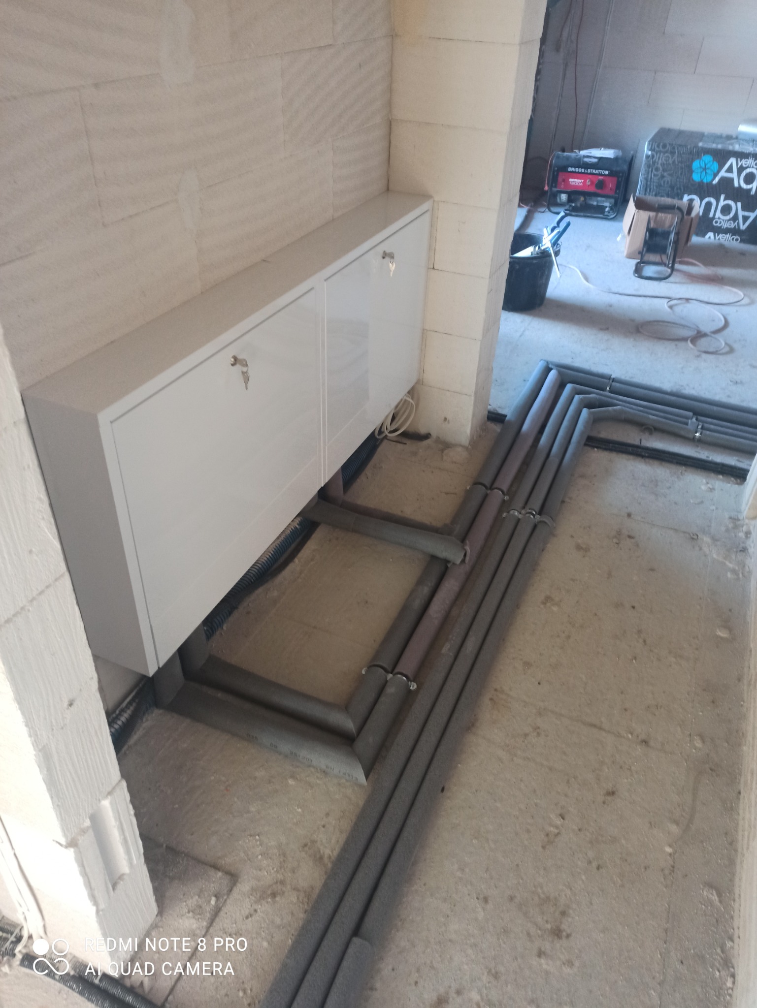 Biała szafka wisząca z kluczykami w nowym budynku, z widocznymi rurami instalacyjnymi zabezpieczonymi izolacją, generator prądu i materiały budowlane w tle.