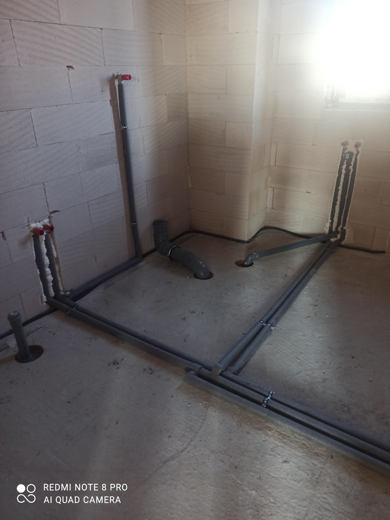 Szare rury kanalizacyjne ułożone na surowym betonowym podłożu w pomieszczeniu z białych bloczków, widoczne pionowe wyprowadzenia ze ścian.