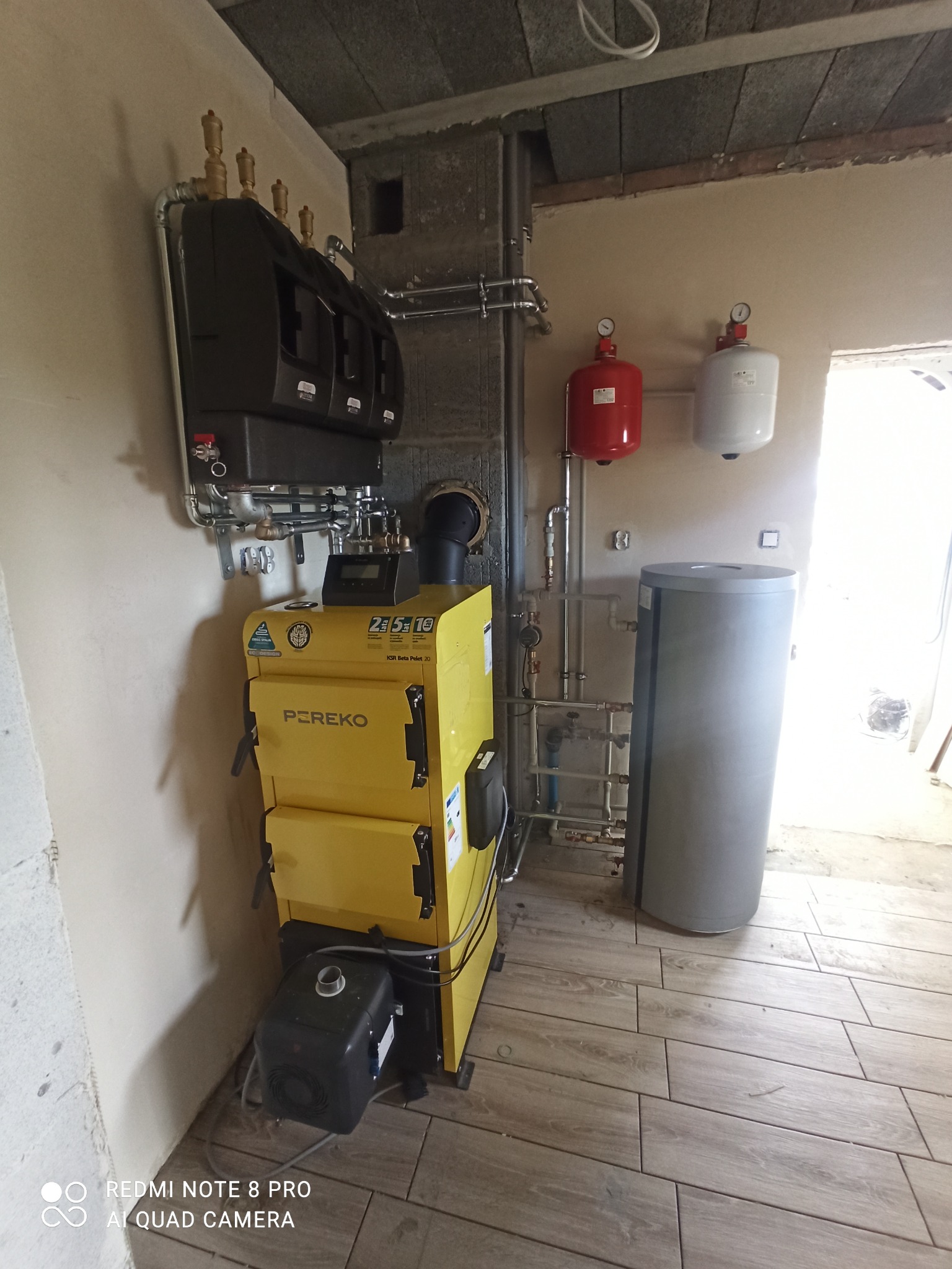 Żółty piec na pellet marki PEREKO KSR Beta Pelet 20, z podłączonymi rurami i zbiornikiem, w trakcie montażu w surowym pomieszczeniu z betonowym sufitem i wykończoną podłogą z płytek imitujących...